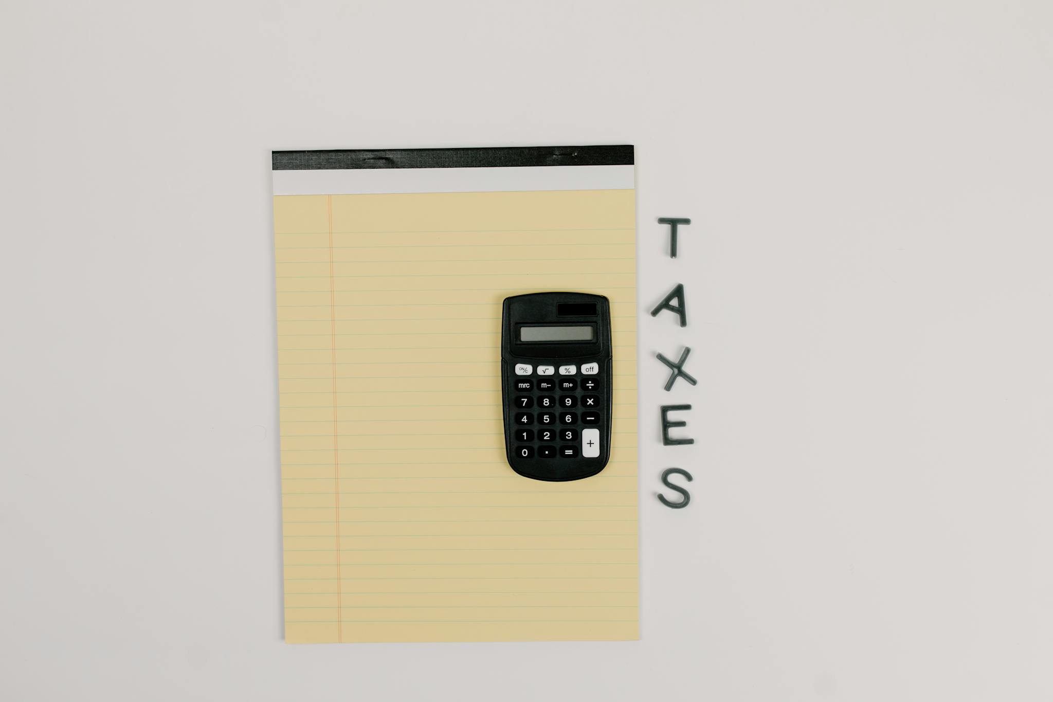 Top-down view of a calculator on a yellow notepad with 'TAXES' spelled out beside.